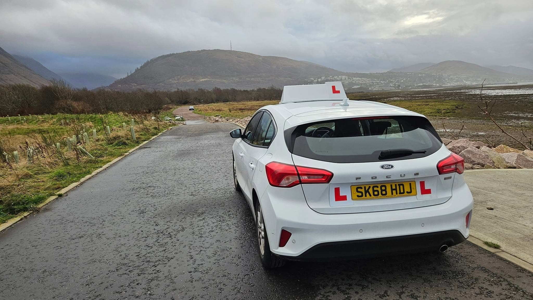LearnWithLeo's white dual-control learner car with L plates on a shoreside road near Fort William, hills in the distance